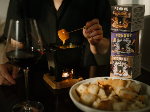 Person dipping fondue into a fondue pot with wine and bread nearby, surrounded by fondue boxes.